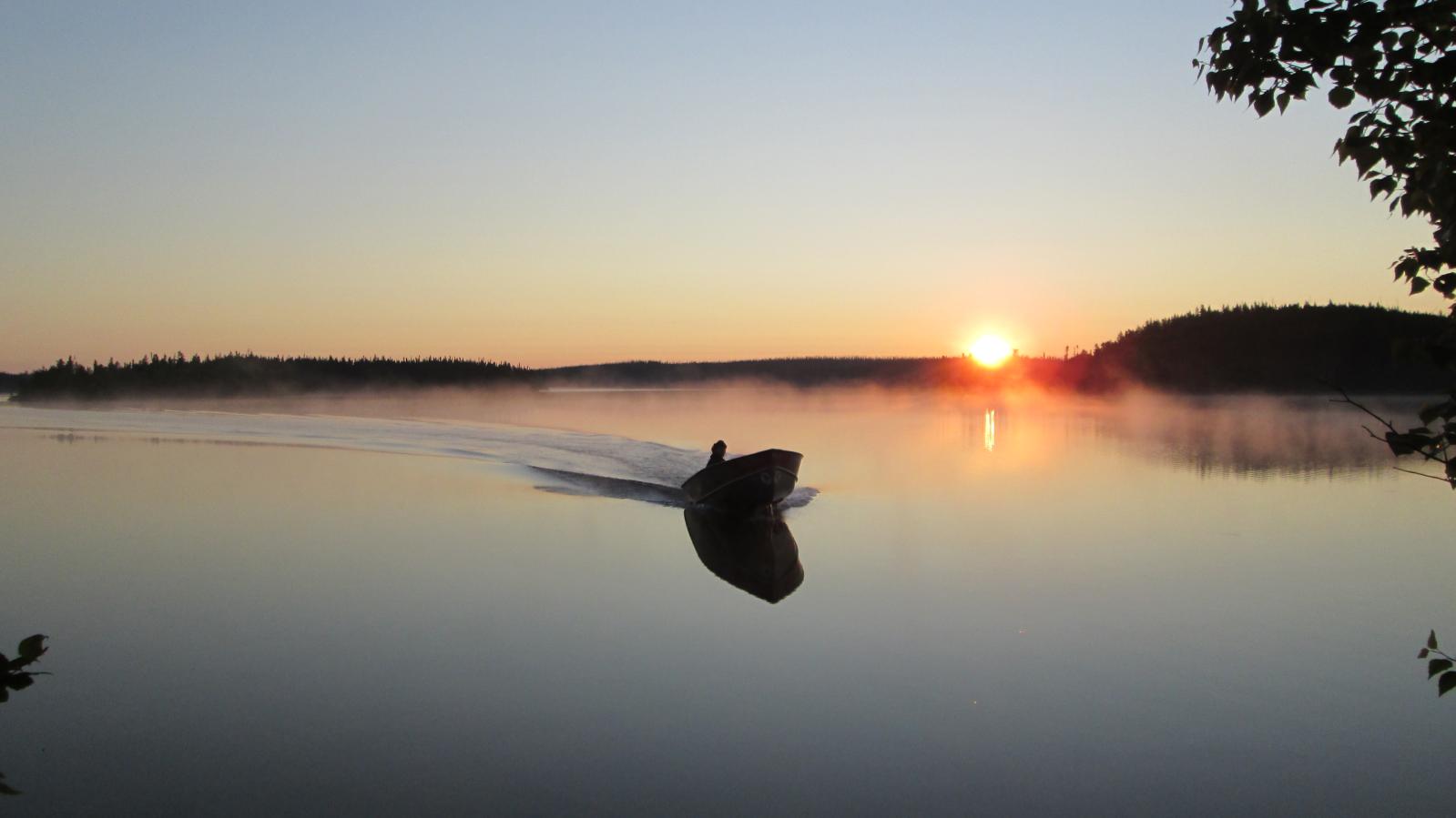 A motor boat returning to camp on a placid lake near dusk with the sun setting in the background 